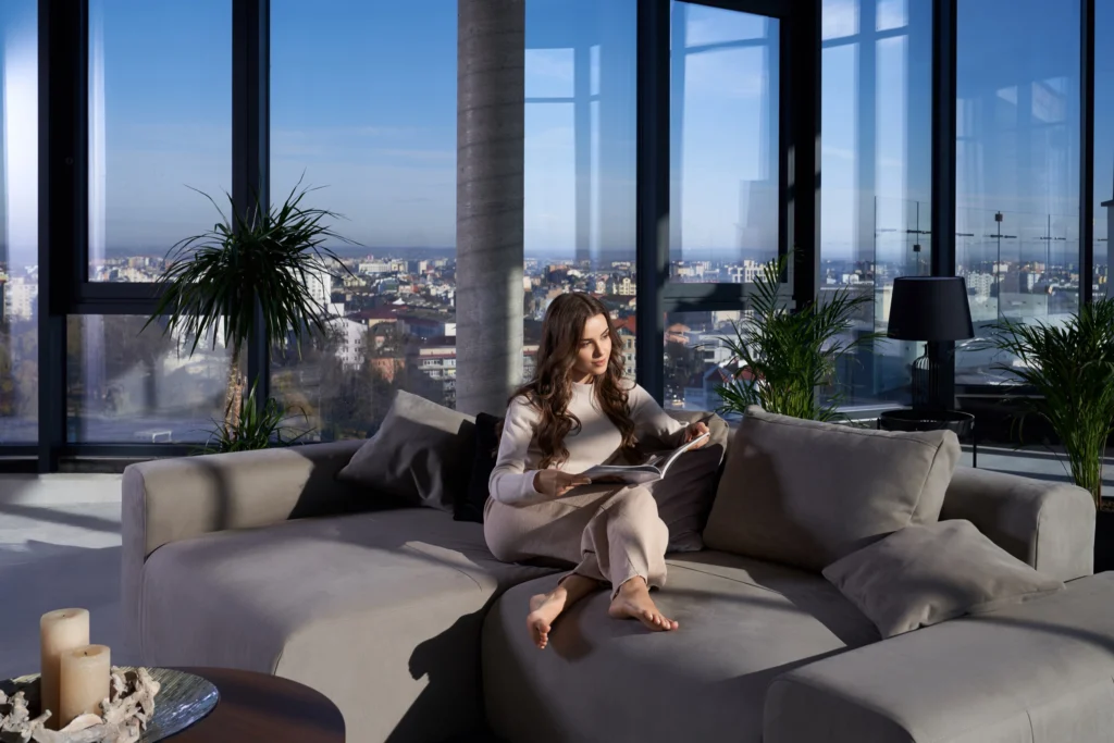 Front view of beautiful brunette woman sitting on grey sofa and reading book or magazine near large panoramic window with incredible view. Concept of resting at home or hotel.