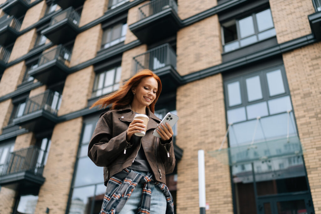 Low-angle view of stylish cheerful young woman standing on city street while drinking coffee and browsing smartphone with smile on face, posing on blurred background of modern building.