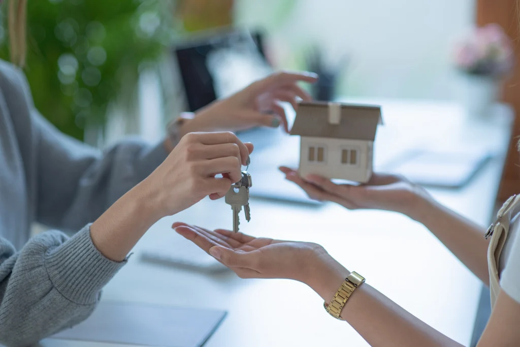 Midsection Of Real Estate Agent Giving House Key To Customer While Sitting In Office