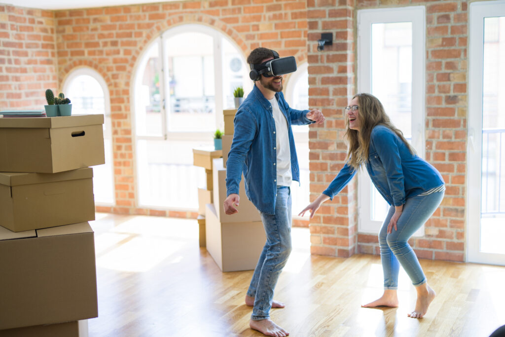 Young couple moving to a new house having fun playing with virtual reality glasses around cardboard boxes
