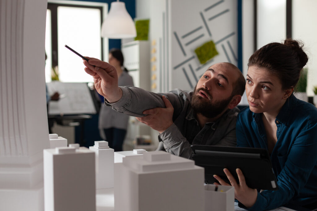 Team of construction architects work on tablet at office to plan building model maquette. Caucasian engineering partners pointing at layout project to design modern digital blueprint