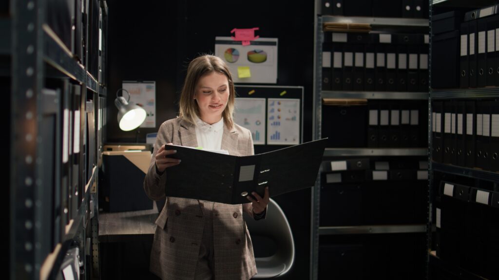 In workplace, confident caucasian detective inspector holds files and evidence, ready to solve crimes. Female law enforcement agent examines case records, smiling and looking at camera. Zoom-in shot.