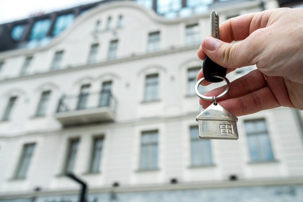 closeup Real estate agent holding key near house outdoors, high-rise buildings of modern city, buy home or renting property