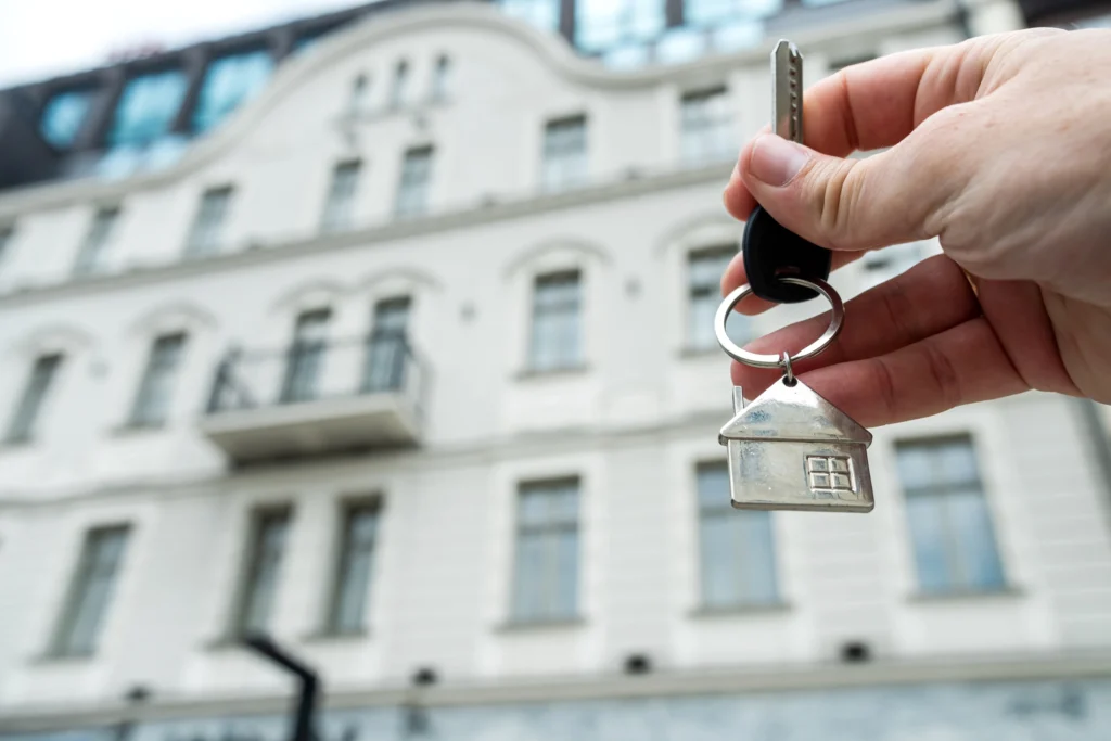 closeup Real estate agent holding key near house outdoors, high-rise buildings of modern city, buy home or renting property