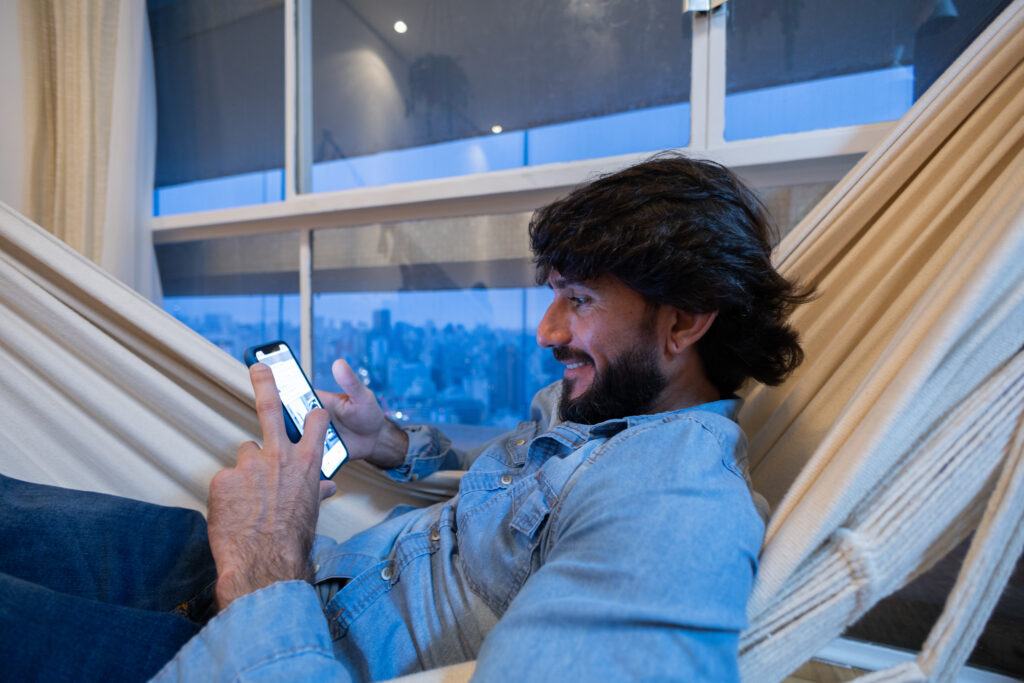 Side view of young man using a smartphone at night time in his living room on a hammock with city view landscape in the background. Mobile phone, technology, concept. Texting on the phone. High quality photo
